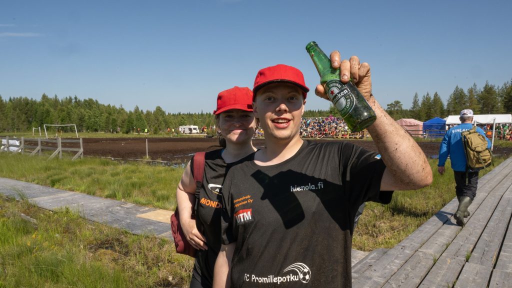 Kaksi nuorta naista katsovat kameraa ja hymyilevät. Toisella naisella on kädessää on olut. Taustalla näkyy Suopotkupallokenttä ja katsomo. Kyseessä on Suopotkupallon MM-kilpailut vuonna 2025