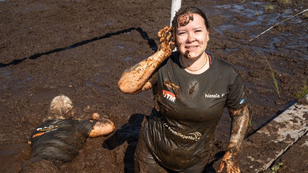 Kuvassa näkyy kaksi naista. Toinen on uponnut suohon ja toinen seisoo ja katsoo kameraa hymyillen, hänkin on yltä päältä mudan peitossa. Kyseessä on Suopotkupallon MM-kilpailut vuonna 2025
