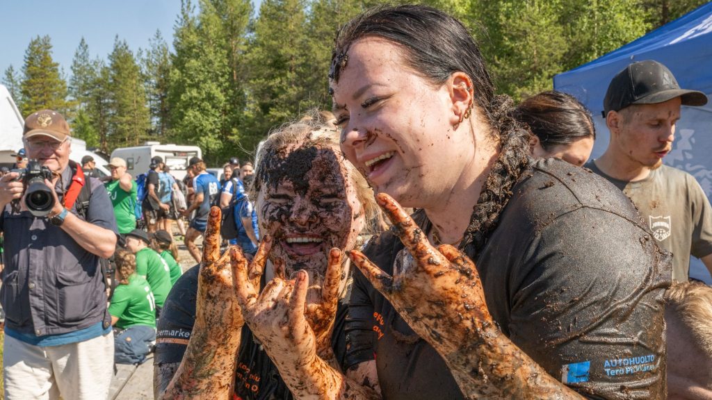 Kaksi yltä päältä mutaista naista nauravat ja näyttävät käsimerkkiä, jossa etusormi ja pikkurilli ovat pystyssä. Kyseessä on Suopotkupallon MM-kilpailut vuonna 2025