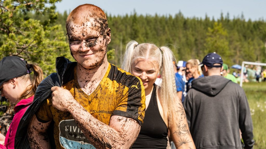 Mutainen mies katsoo kameraa ja hymyilee. Taustalla näkyy muitakin ihmisiä. Kyseessä on Suopotkupallon MM-kilpailut vuonna 2025