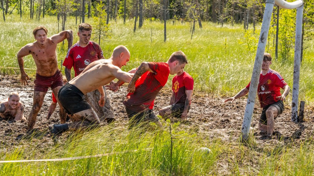 Suomalainen joukkue pelaa Englantilaista joukkuetta vastaan suopotkupalloa. Iso-Britannian joukkueella on punaiset pelipaidat, missä lukee manchester united. Kyseessä on Suopotkupallon MM-kilpailut vuonna 2025. (In Enlish: Finnish and Brittish teams are playing Swamp Soccer. British players have red shirts that say manchester united. They are in Swamp SOccer World Championships 2025