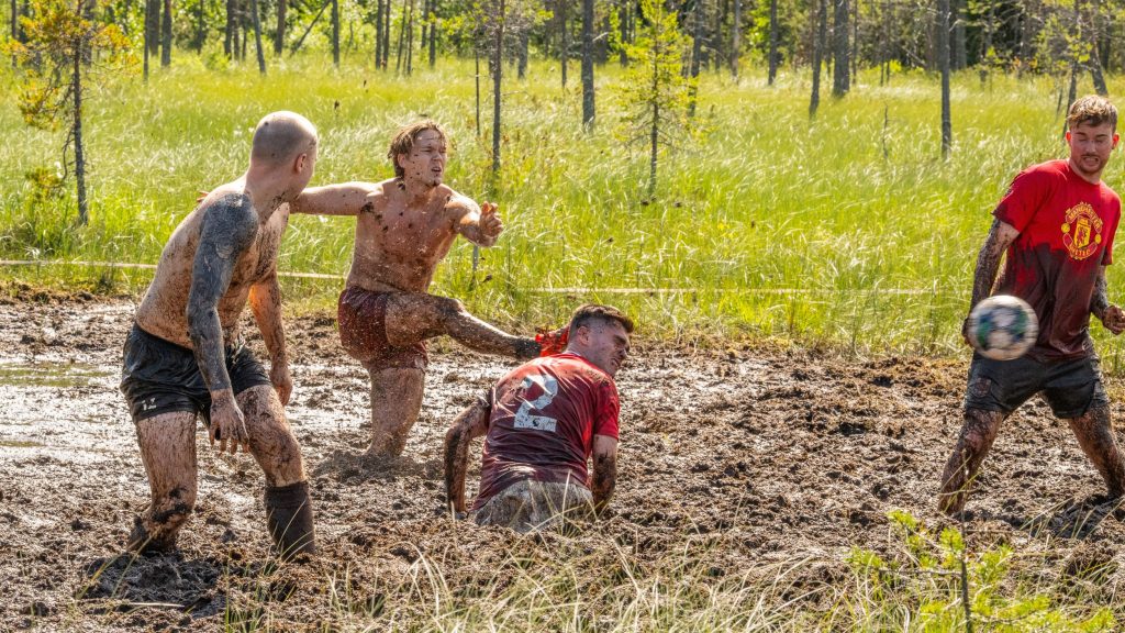 Suomalainen joukkue pelaa Englantilaista joukkuetta vastaan suopotkupalloa. Iso-Britannian joukkueella on punaiset pelipaidat, missä lukee manchester united. Kyseessä on Suopotkupallon MM-kilpailut vuonna 2025. (In Enlish: Finnish and Brittish teams are playing Swamp Soccer. British players have red shirts that say manchester united. They are in Swamp SOccer World Championships 2025