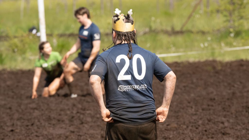 Mies seisoo selin kameraan päin. Hänellä on kruunu päässään. Taustalla näkyy kaksi muutakin ihmistä. Kyseessä on Suopotkupallon MM-kisat 2025