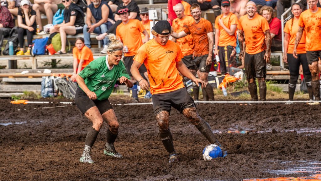 Mies ja nainen pelaavat jalkapalloa suolla. Taustalla yleisöä. Kyseessä on Suopotkupallon MM-kisat 2025