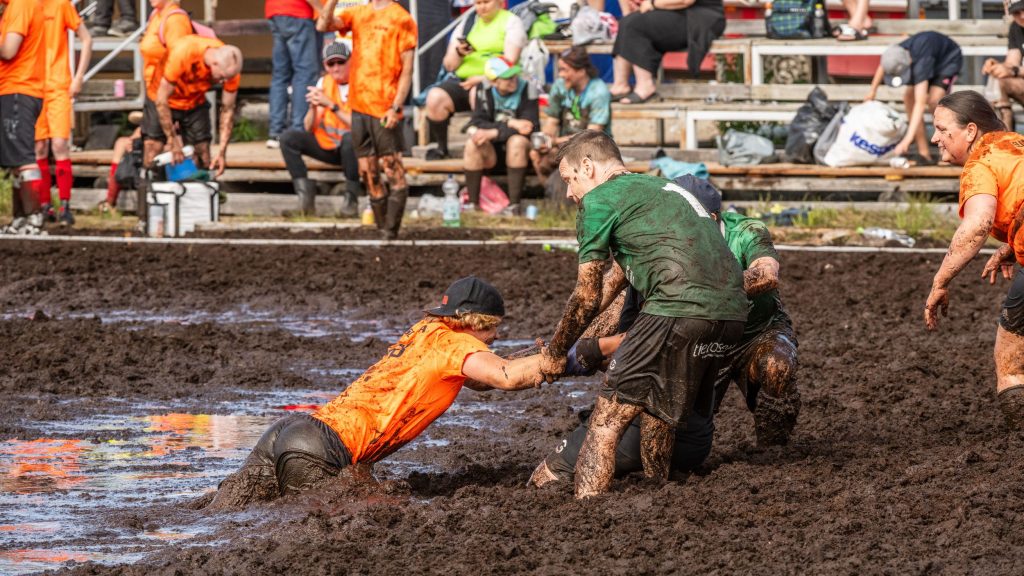 Miehet vetävät toista miestä pois suosta. Taustalla on yleisöä. Kyseessä on Suopotkupallon MM-kisat 2025