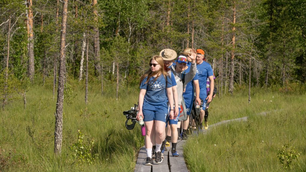 Sinisiin joukkuepaitoihin pukeutuneeteet Suopotkupallon pelaajat kävelevät jonossa pitkospuita pitkin. Yksi pelaajista katsoo kameraa hymyillen ja vilkuttaa. Kyseessä on Suopotkupallon MM-kilpailut vuonna 2025
