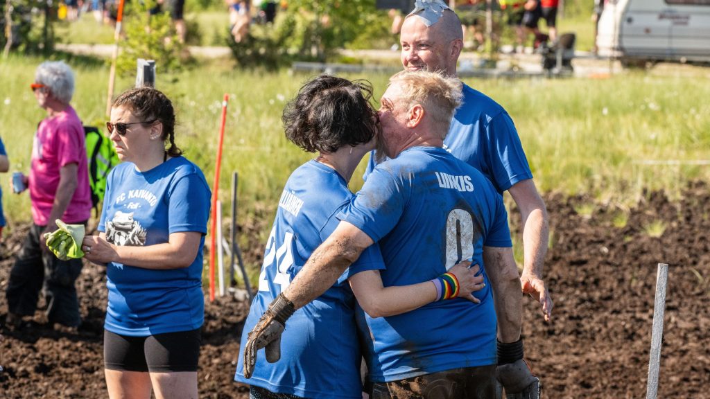 Pelipaitoihin pukeutuneet keski-ikäiset mies ja nainen pussaavat seisoessaan suolla. Kyseessä on Suopotkupallon MM-kisat 2025