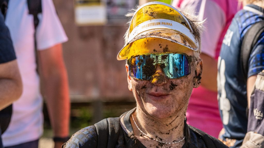Yltä päältä mudan peitossa oleva aurinkolasipäinen mies katsoo kameraa ja hymyilee. Kyseessä on Suopotkupallon MM-kisat 2025