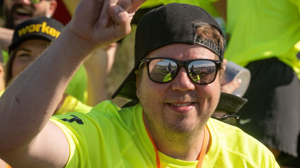 Aurinkolasipäinen mies katsoo kameraa hymyillen ja tuulettaa toisella kädellään. Kyseessä on Suopotkupallon MM-kilpailut vuonna 2025