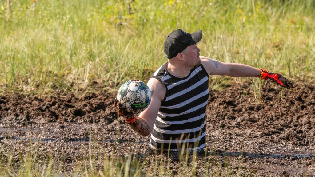 Mies heittää jalkapalloa suolla. Hän on uponnut vyötäröön asti suohon. Kyseessä on Suopotkupallon MM-kisat 2025