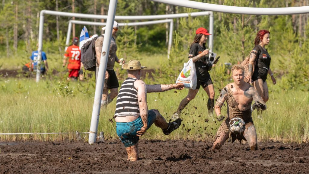 Mies potkaisee jalkapalloa maalia kohti ja maalivahti yrittää torjua palloa. He pelaavat suolla. Kyseessä on Suopotkupallon MM-kilpailut vuonna 2025