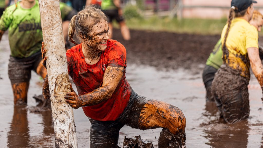 Nainen nojaa maalin tolppaan ja koittaa nousta upottavasta suosta. Hän hymyilee. Kyseessä on suopotkupallon MM-kisat 2024