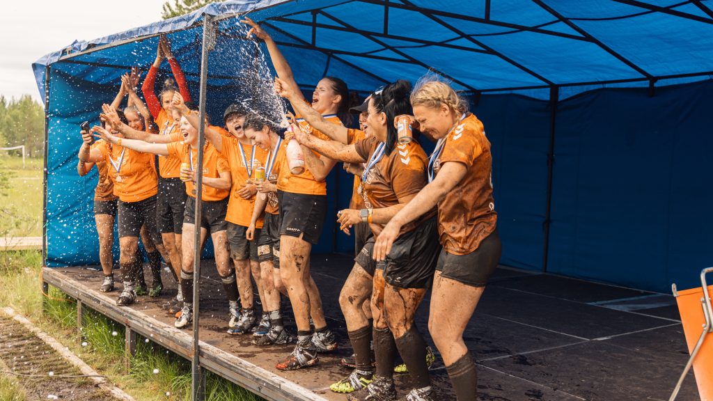 Naiset seisovat esiintymislavalla tuulettaen ja juhlien. Yksi suihkuttaa shampanjaa/kuohuviiniä. Kyseessä on suopotkupallon MM-kisojen 2024 mitalien jako